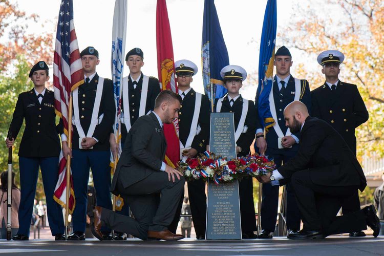 Men placing wreath around a marker, with ROTC students holding flags in background.