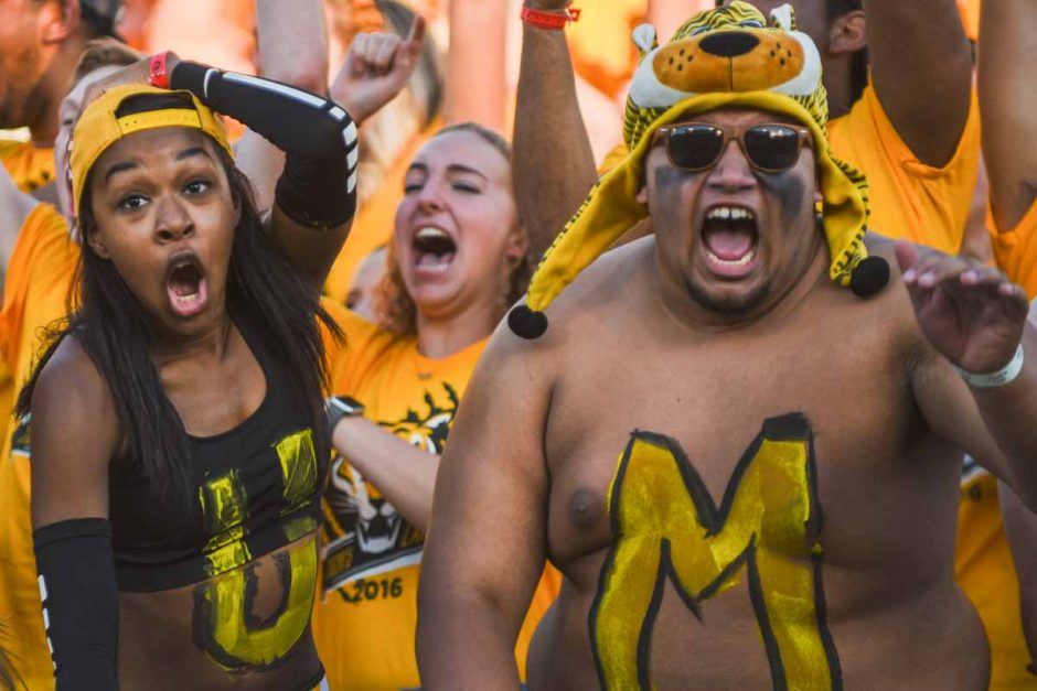 Students in body paint cheering in the bleachers at a football game.
