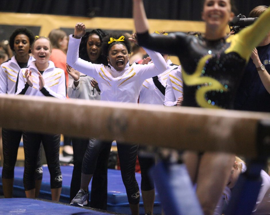 Gymnasts in warmup suits cheer as a teammate dismounts from the beam.