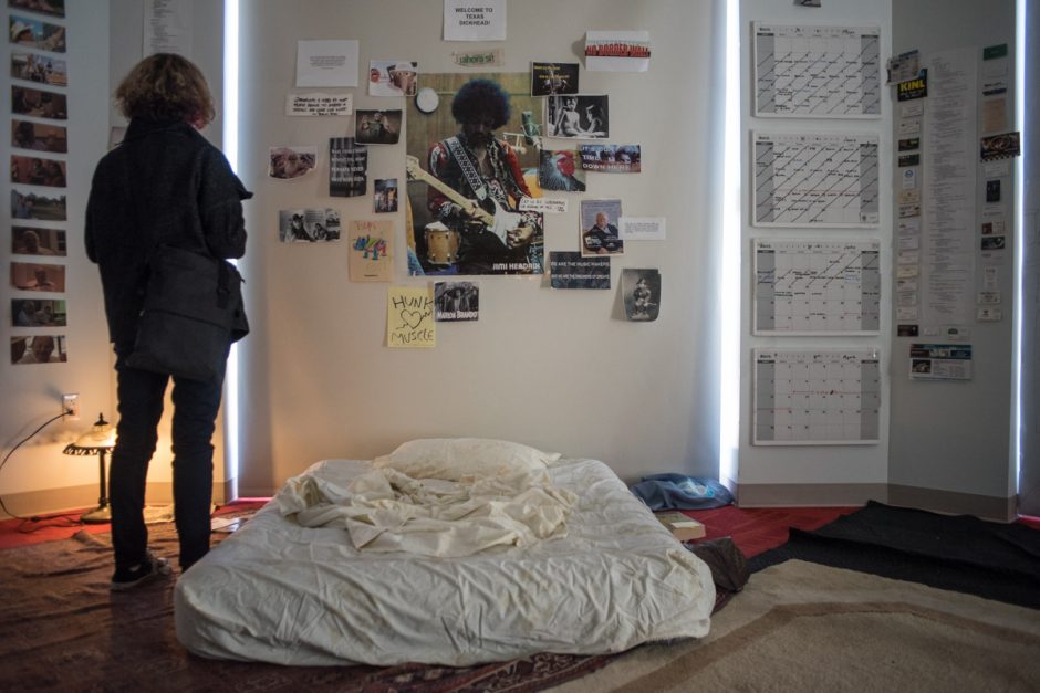 Photos of Jimi Hendrix, calendars and other papers on a wall over a mattress.