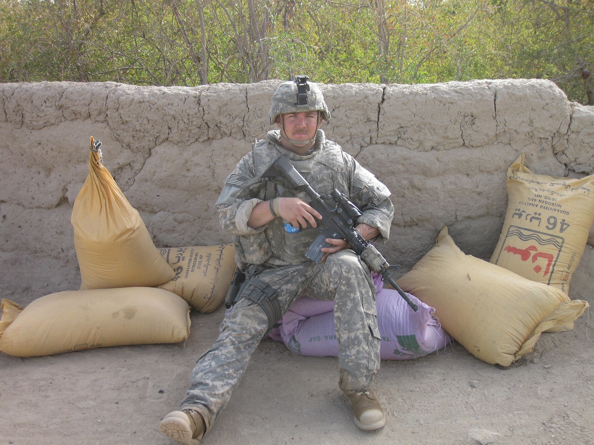 Alex Pracht, dressed in a military uniform and holding a weapon, sitting on the ground in front of wall.